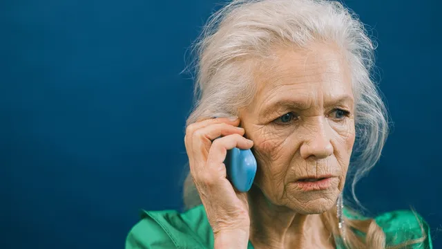 Mulher idosa de cabelo branco e blusa verde fala ao telemóvel com ar preocupado, sobre um fundo azul