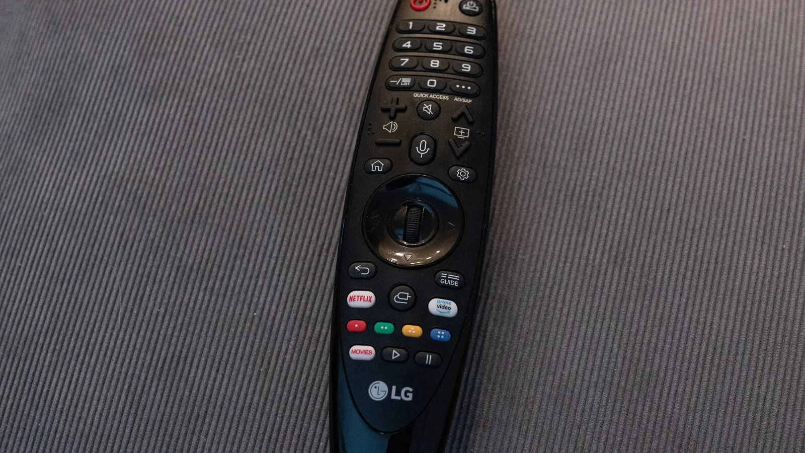 Comando Magic Remote da LG, preto, pousado num sofá cinzento, com botões de volume, microfone, definições e atalhos para Netflix e Prime Video