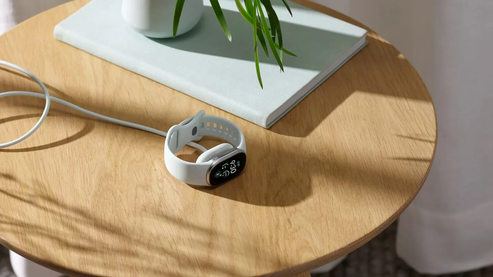 Pulseira Fitbit Edge branca a carregar sobre uma mesa redonda de madeira, ao lado de um livro e de uma planta