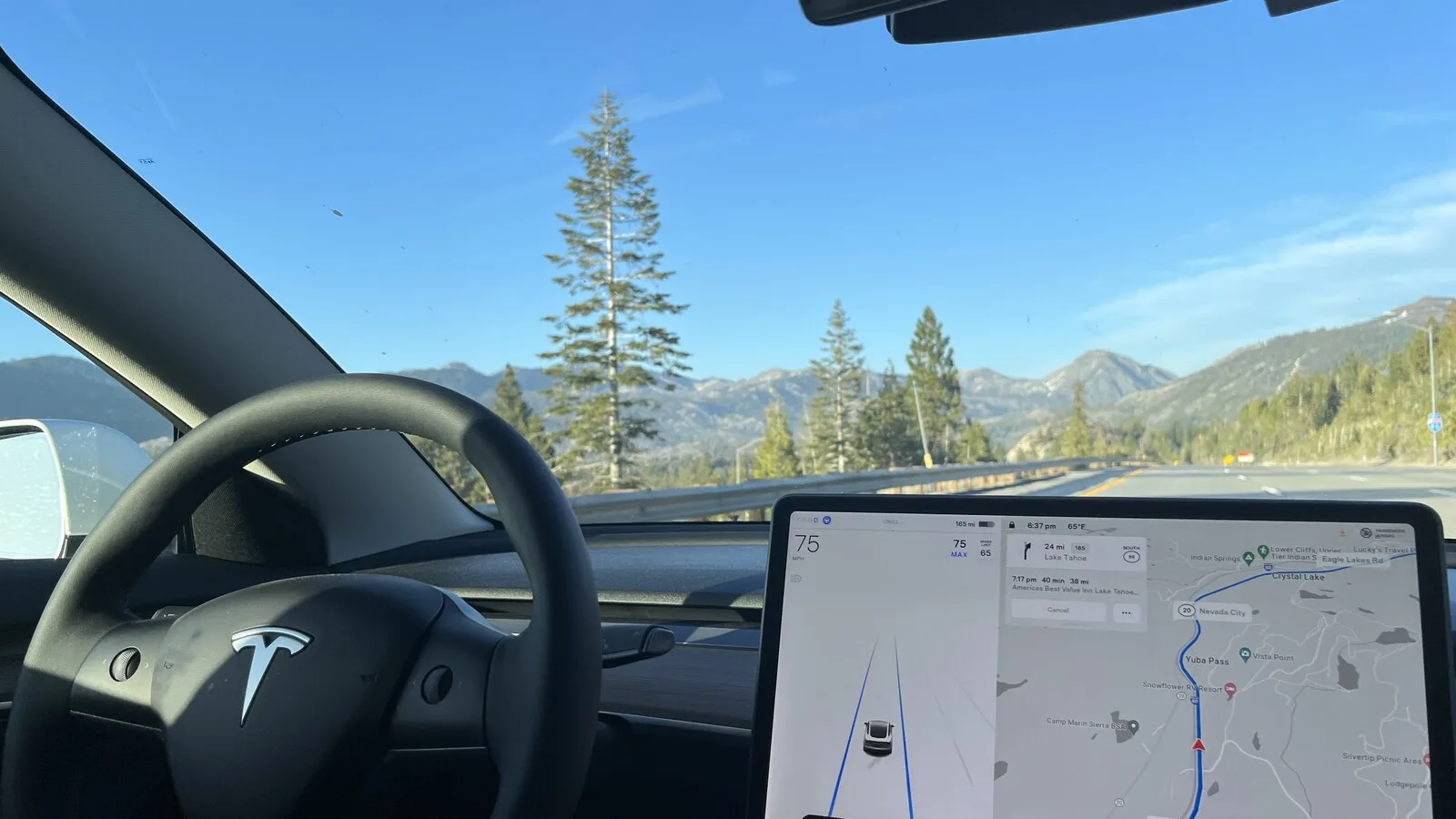 Interior de um Tesla numa autoestrada de montanha, com o volante à esquerda e o ecrã central a mostrar o carro na faixa e um mapa