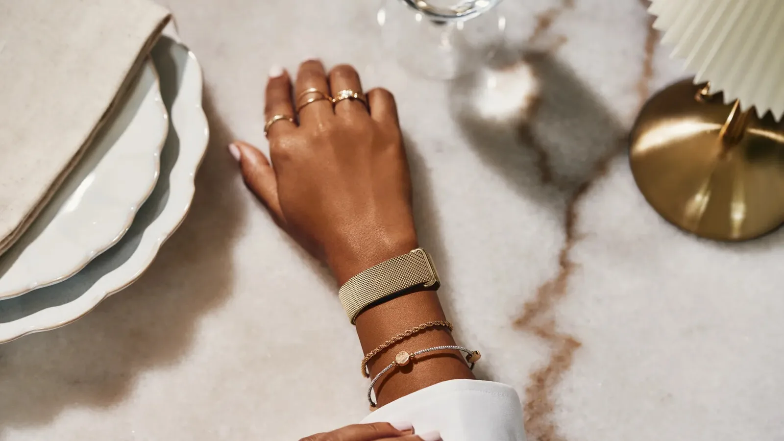 Mão de uma mulher pousada numa mesa de mármore, com a Fitbit Air presa ao pulso por uma bracelete de malha metálica dourada, ao lado de pulseiras e de pratos