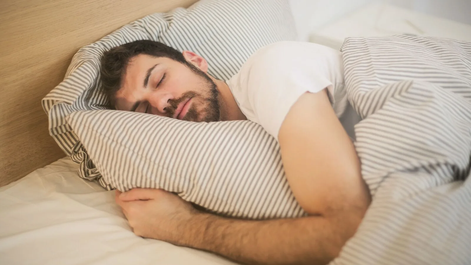 Homem de barba a dormir de lado numa cama, abraçado a uma almofada com fronha às riscas cinzentas e brancas