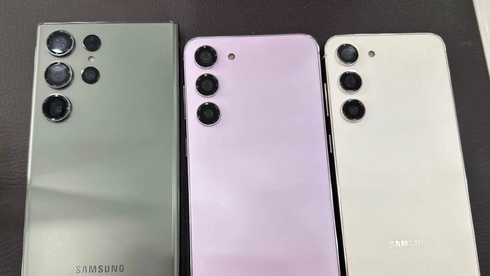 Traseiras de três telemóveis da série Galaxy S23, em verde, lilás e creme, lado a lado sobre uma superfície escura, com as câmaras verticais à vista