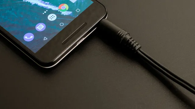 Parte inferior de um telemóvel Android preto pousado numa superfície escura, com o ecrã inicial ligado e um cabo preto encaixado