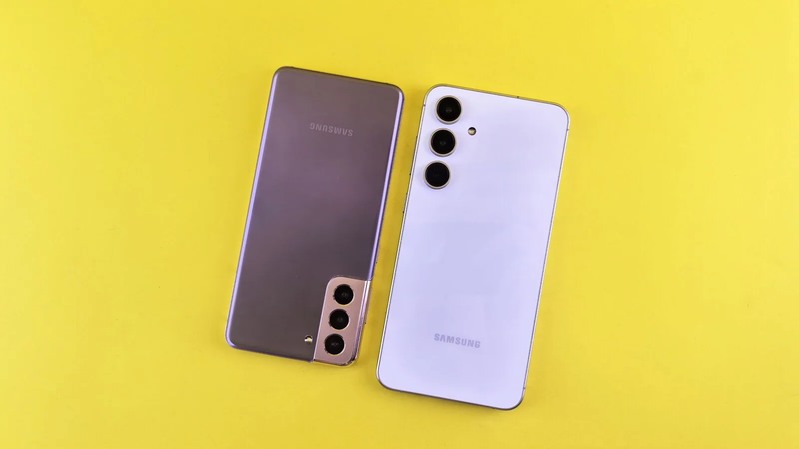 Dois telemóveis Samsung Galaxy vistos de trás, um lilás e outro branco, pousados lado a lado sobre um fundo amarelo