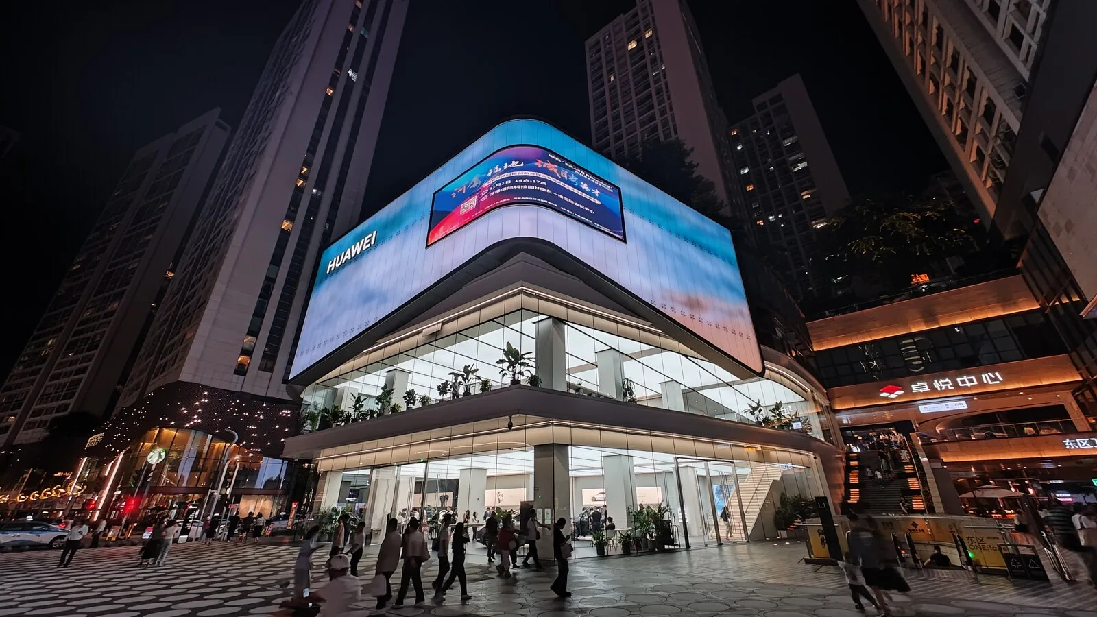 Loja da Huawei num centro comercial de Shenzhen, à noite, com fachada de vidro iluminada e um grande ecrã curvo no topo, e pessoas a passar na praça em frente