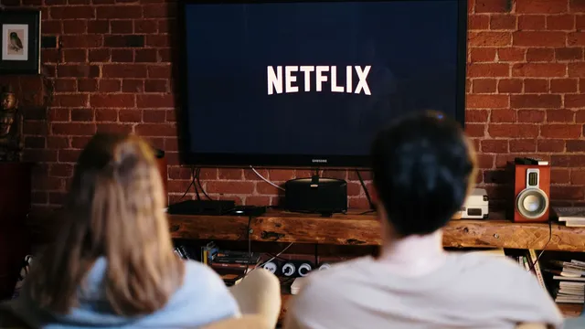Duas pessoas de costas, sentadas em frente a um televisor com o logótipo da Netflix numa sala com parede de tijolo