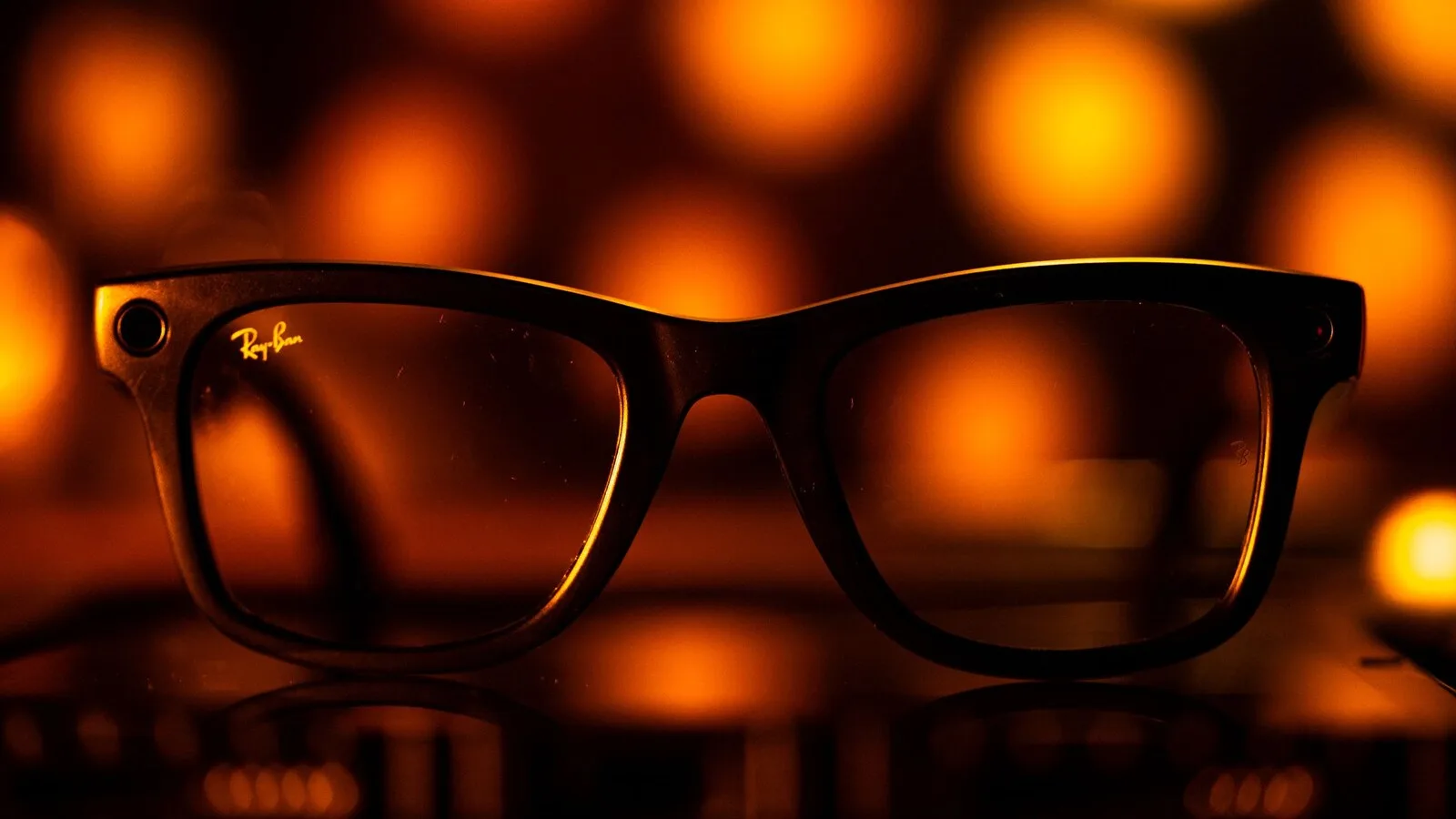 Óculos Ray-Ban Meta pretos pousados numa superfície escura, com a pequena câmara visível no canto da armação e luzes alaranjadas desfocadas ao fundo