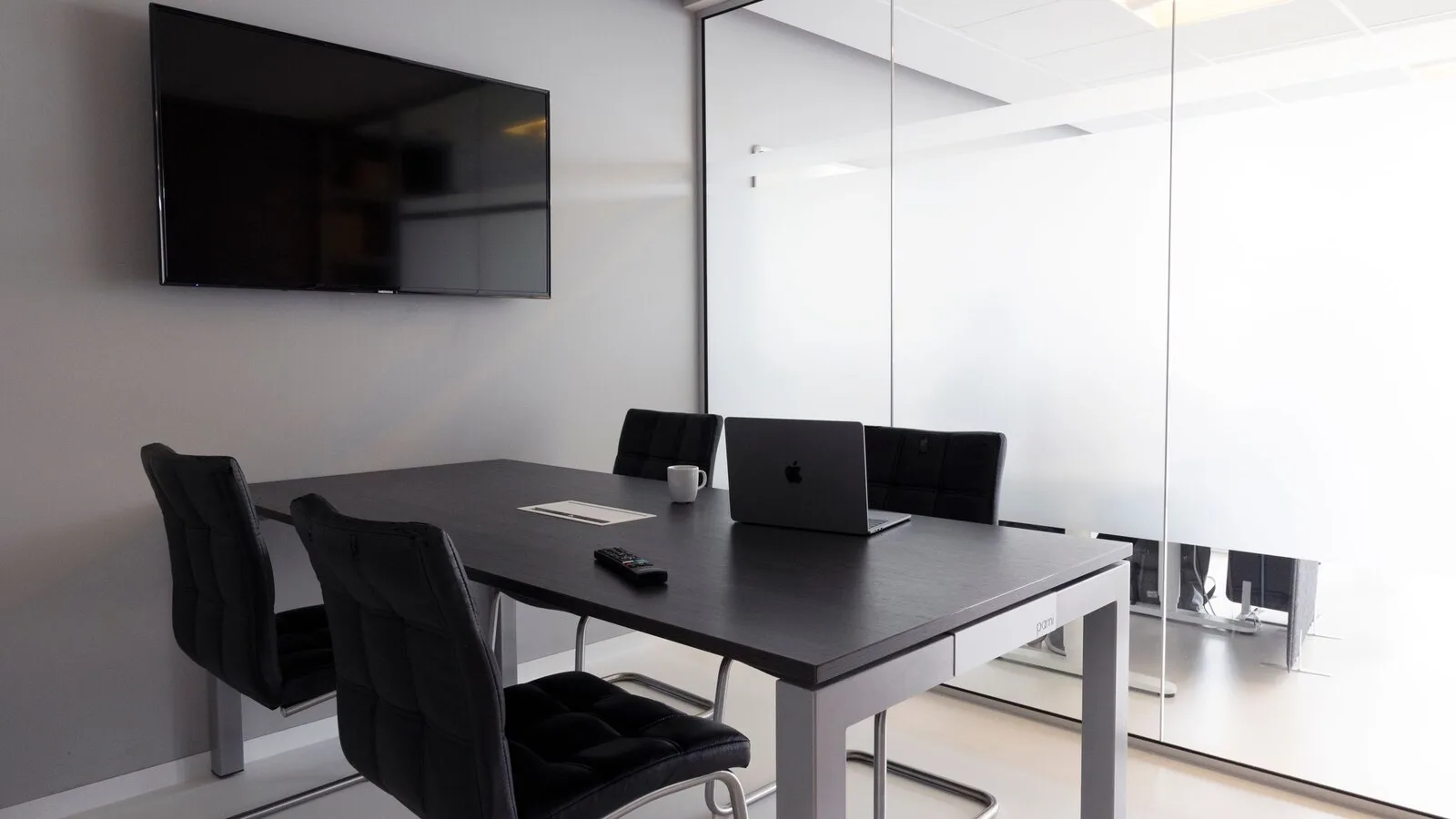 Sala de reuniões vazia com mesa escura, cadeiras pretas, um portátil e um televisor na parede