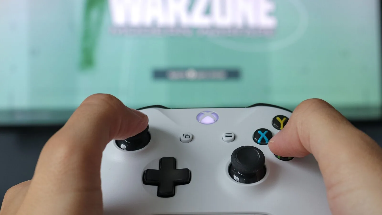 Mãos a segurar um comando Xbox branco em frente a um televisor desfocado com um jogo no ecrã