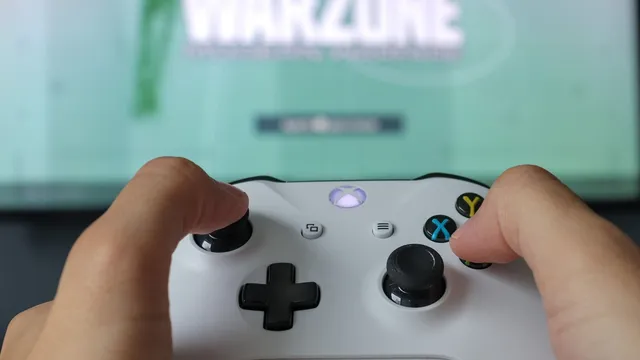 Mãos a segurar um comando Xbox branco em frente a um televisor desfocado com um jogo no ecrã
