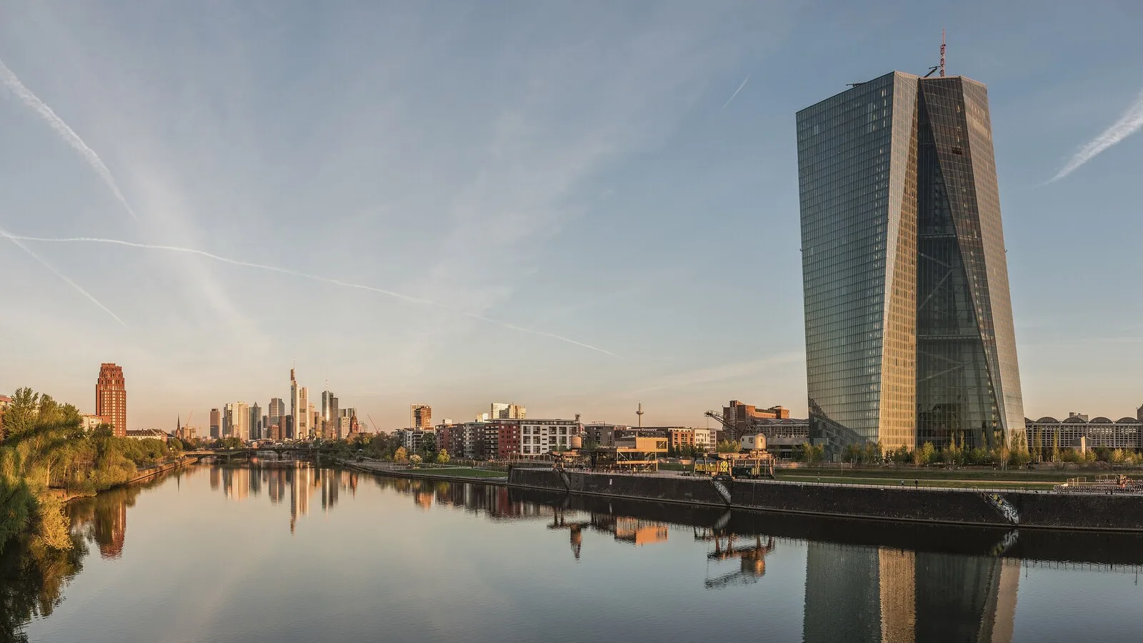 Torre de vidro da sede do Banco Central Europeu junto ao rio Meno, em Frankfurt, com os arranha-céus da cidade ao fundo, ao amanhecer