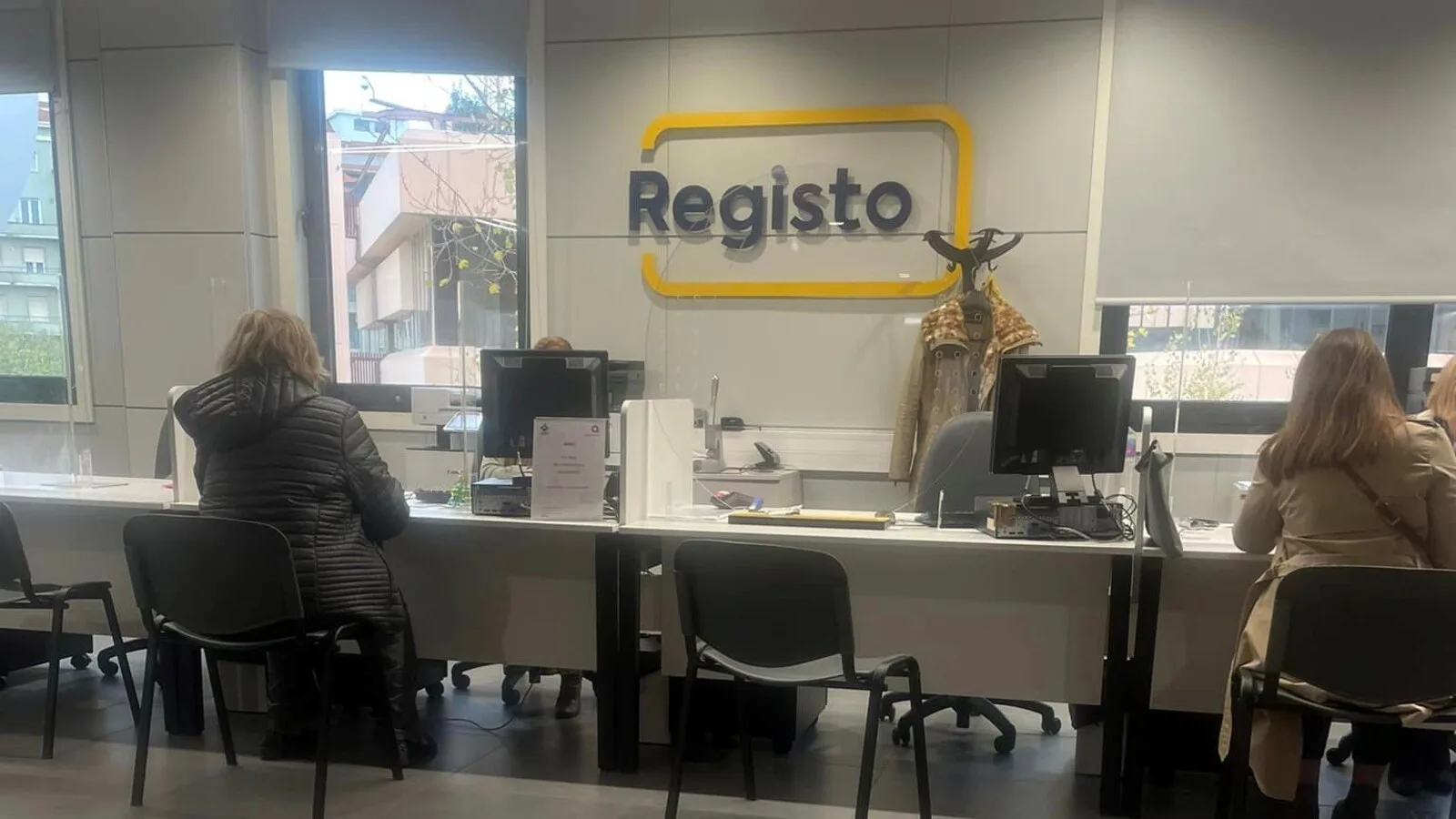 Duas pessoas sentadas de costas ao balcão do Registo numa Loja de Cidadão, com monitores e um letreiro amarelo com a palavra Registo na parede