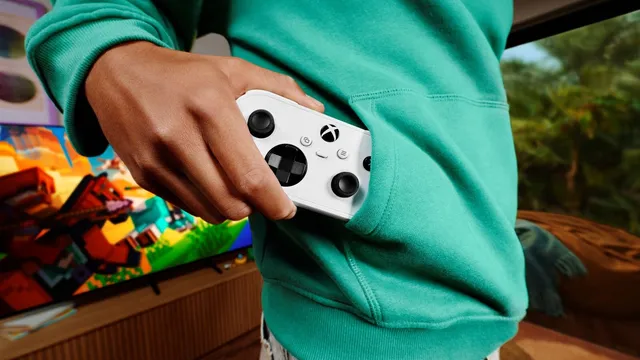 Mão a guardar um pequeno comando Xbox branco no bolso de uma camisola verde, com um jogo de blocos coloridos num televisor ao fundo