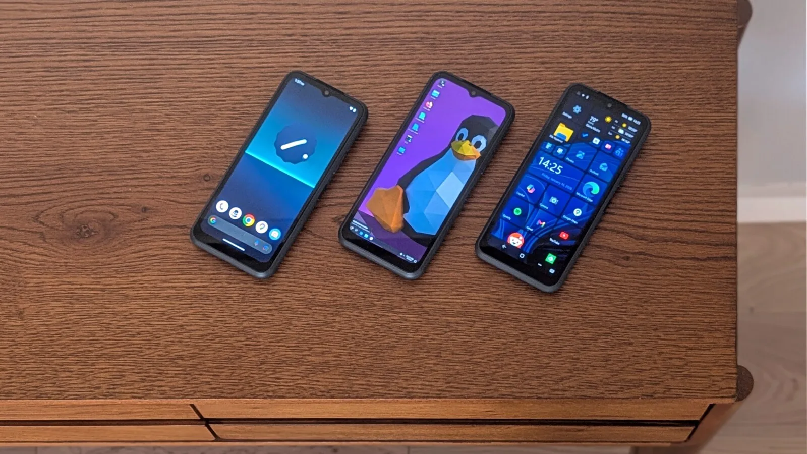 Três telemóveis NexPhone pousados numa mesa de madeira, um com o ecrã do Android, outro com o pinguim do Linux e outro com mosaicos do Windows