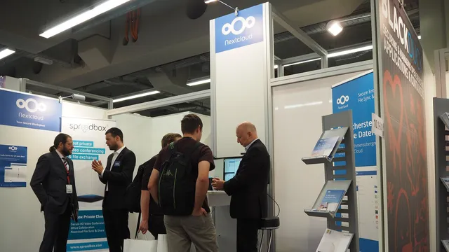 Visitantes junto ao expositor do Nextcloud numa feira de tecnologia, com o logótipo azul da marca no topo de uma coluna