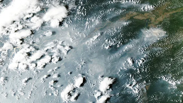 Imagem de satélite da floresta amazónica coberta por fumo azulado e nuvens brancas, com o rio Amazonas a atravessar a imagem