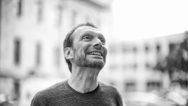 Fotografia a preto e branco de Toni Schneider, sorridente, a olhar para cima numa rua com edifícios desfocados ao fundo