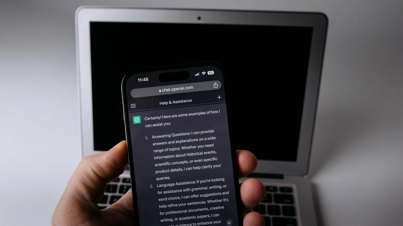 Mão a segurar um telemóvel com uma conversa do ChatGPT aberta no browser, à frente de um portátil com o ecrã desligado