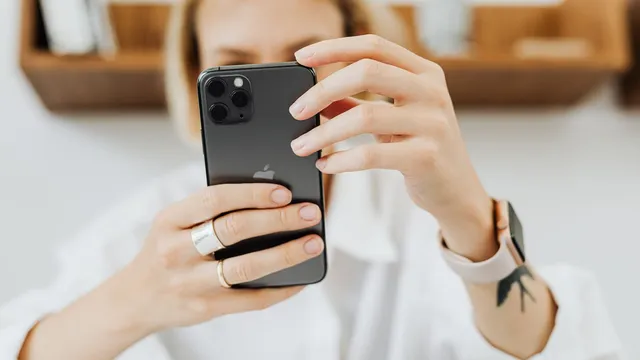 Mulher de camisa branca segura um iPhone à frente da cara para tirar uma fotografia dentro de casa, com uma prateleira de madeira ao fundo