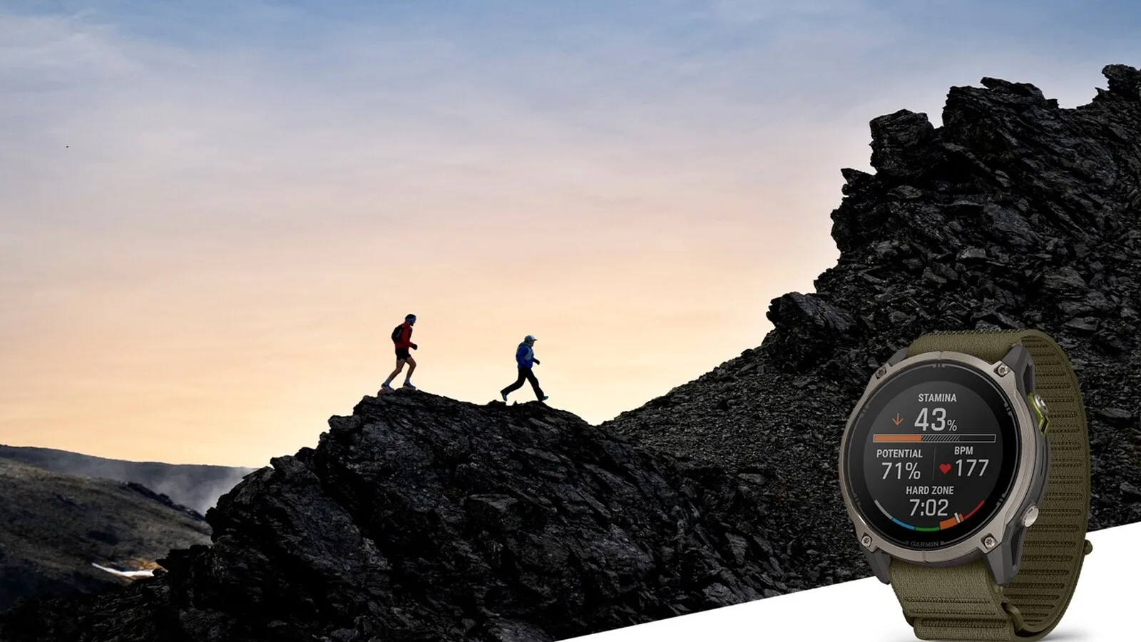 Duas pessoas a correr numa crista rochosa ao entardecer e, em primeiro plano, um relógio Garmin Enduro 4 com bracelete de tecido verde e o ecrã a mostrar a resistência e a frequência cardíaca