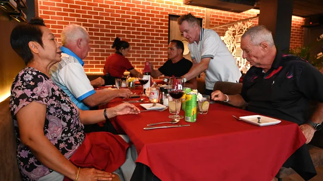 Grupo de pessoas sentadas à mesa de um restaurante com toalha vermelha, copos de vinho e pratos, junto a uma parede de tijolo