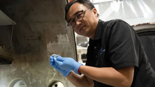 Investigador de óculos e luvas azuis segura uma pequena bateria tipo moeda junto à porta de uma câmara de vácuo térmico, num laboratório