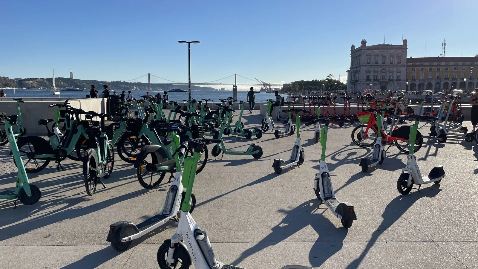 Dezenas de trotinetes e bicicletas elétricas partilhadas estacionadas junto ao Terreiro do Paço, em Lisboa, com o Tejo e a Ponte 25 de Abril ao fundo