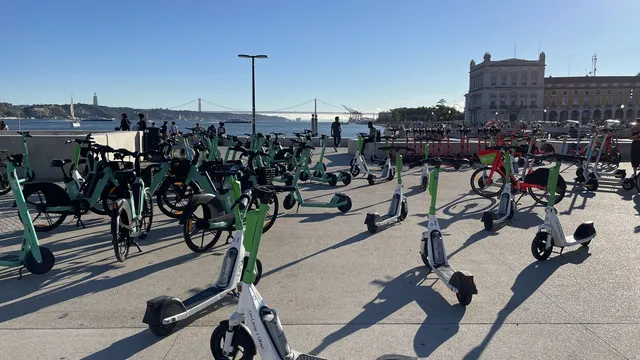 Dezenas de trotinetes e bicicletas elétricas partilhadas estacionadas junto ao Terreiro do Paço, em Lisboa, com o Tejo e a Ponte 25 de Abril ao fundo