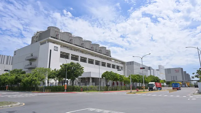 Edifício branco da fábrica Fab 18 da TSMC, com equipamento de refrigeração no telhado, visto de um cruzamento com camiões a passar