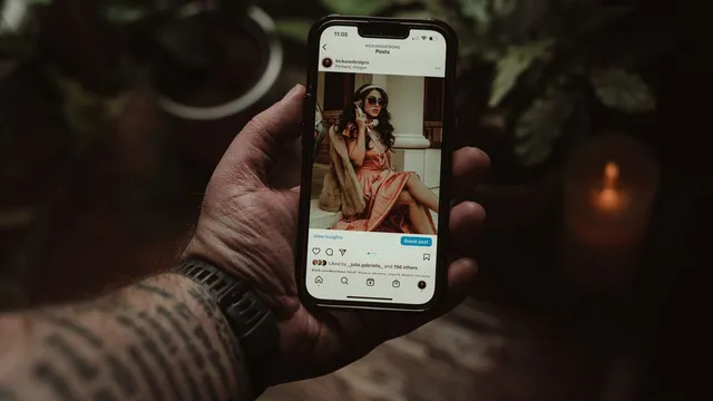Mão tatuada a segurar um iPhone com uma publicação do Instagram aberta, num ambiente escuro com plantas e uma vela ao fundo