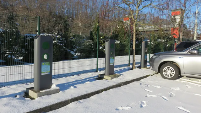 Três postos de carregamento cinzentos num parque coberto de neve, junto a uma vedação verde, com um carro prateado estacionado à direita