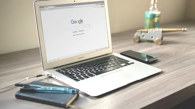Portátil aberto na página da Google em cima de uma secretária de madeira, ao lado de um telemóvel e de um caderno com canetas