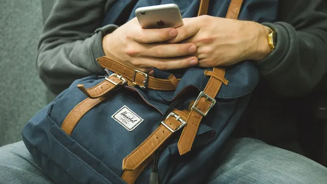 Pessoa sentada com uma mochila azul ao colo a usar um telemóvel com as duas mãos