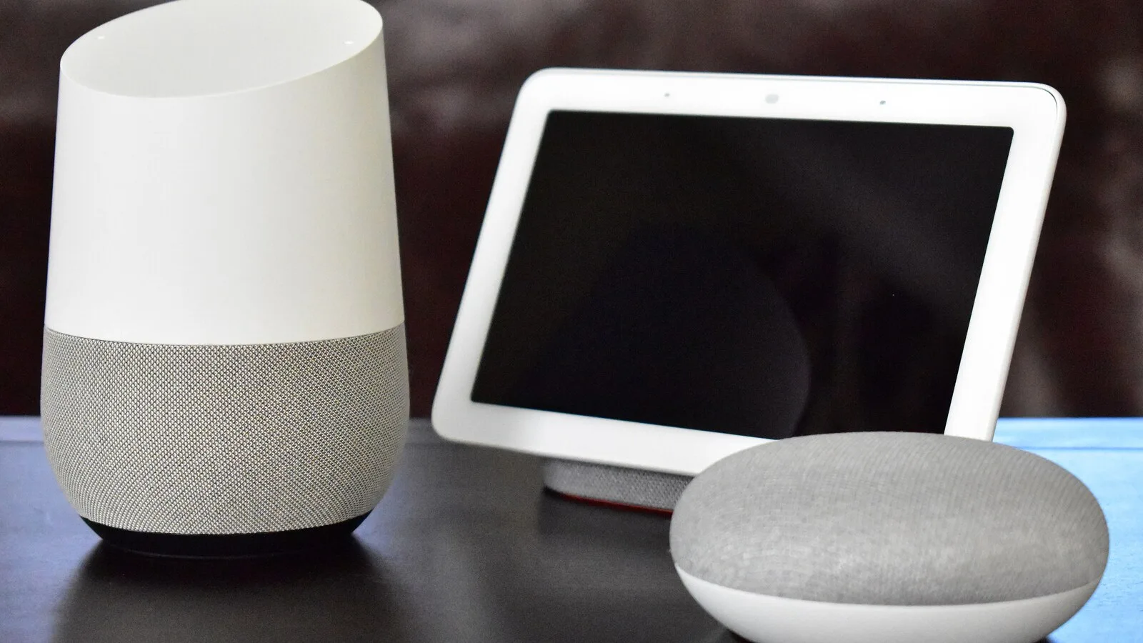 Coluna Google Home branca e cinzenta, ecrã Google Home Hub e coluna redonda Google Home Mini numa mesa escura