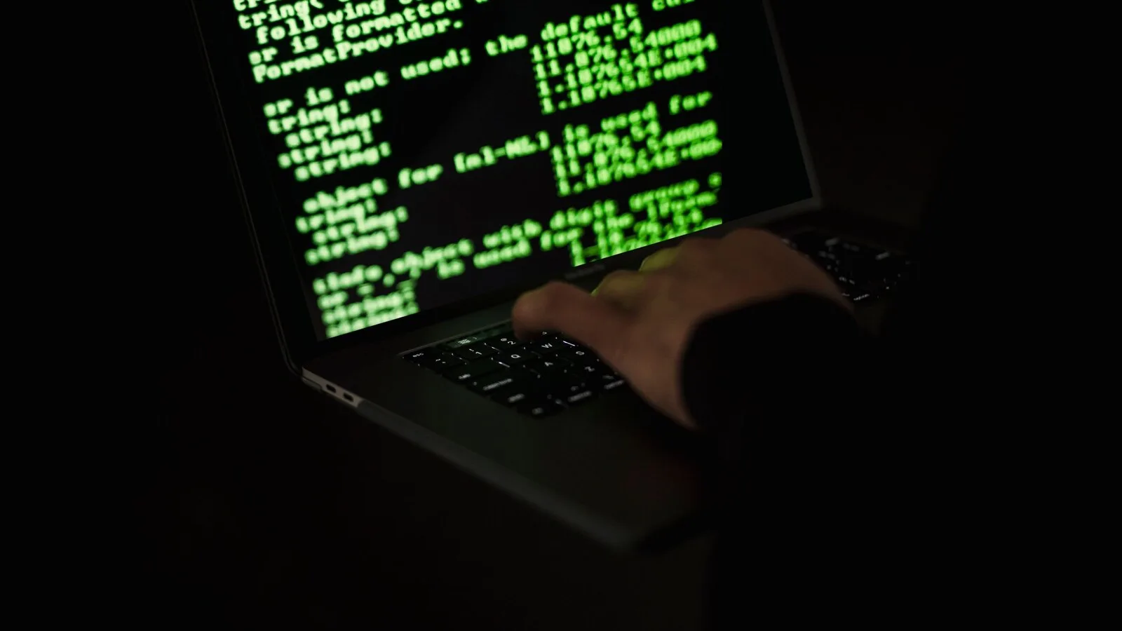 Mão a escrever no teclado de um portátil às escuras, com linhas de código verdes no ecrã