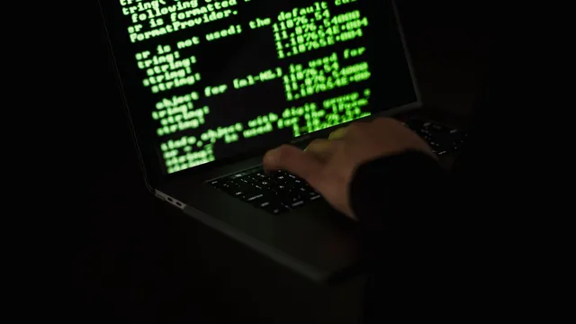 Mão a escrever no teclado de um portátil às escuras, com linhas de código verdes no ecrã