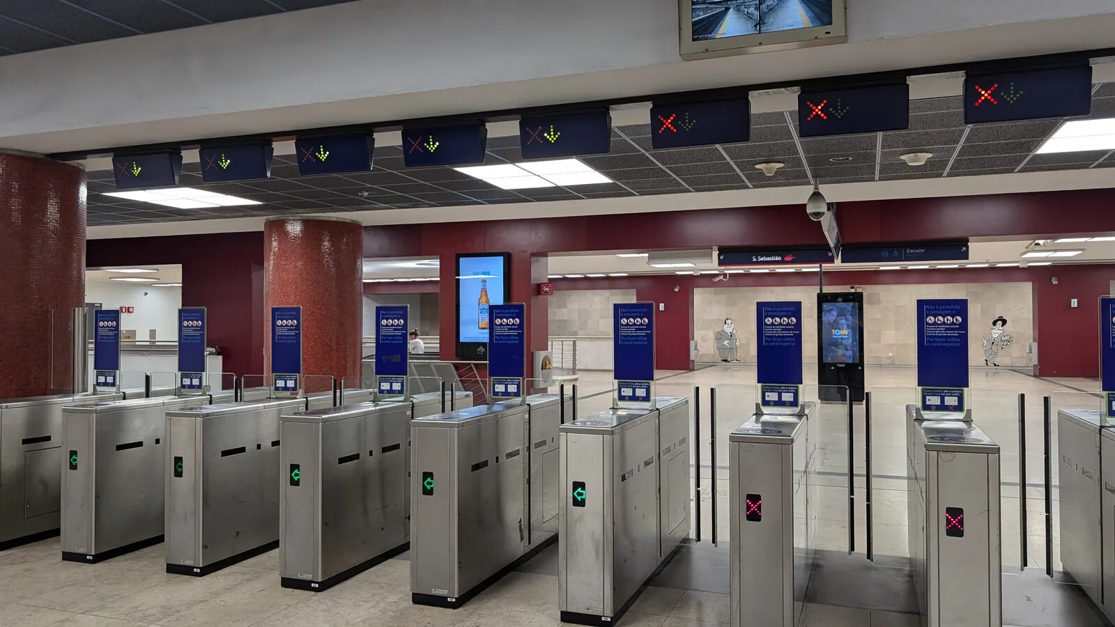 Fila de canais de acesso metálicos numa estação de metro, com setas verdes e cruzes vermelhas por cima e painéis azuis com informação