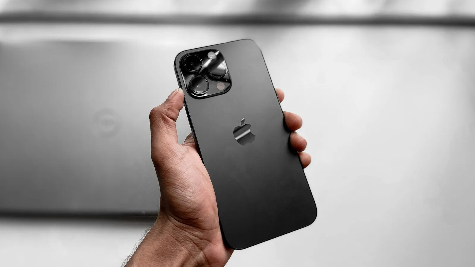 Mão a segurar um iPhone cinzento-escuro de costas, com o módulo de três câmaras e o logótipo da Apple, diante de um fundo claro desfocado