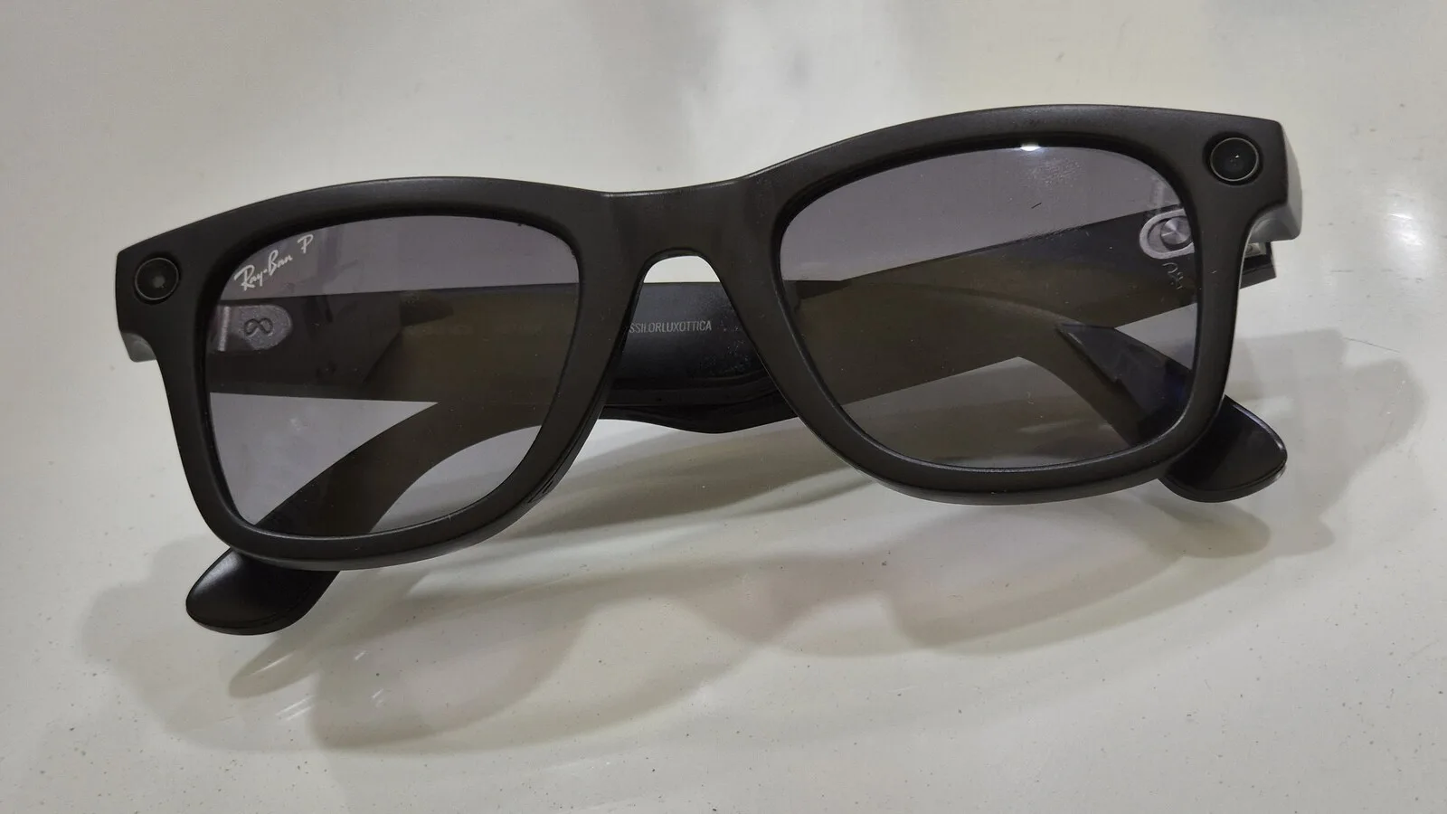 Óculos Ray-Ban Meta Wayfarer pretos com lentes escuras pousados numa mesa branca, com as duas pequenas câmaras visíveis nos cantos da armação