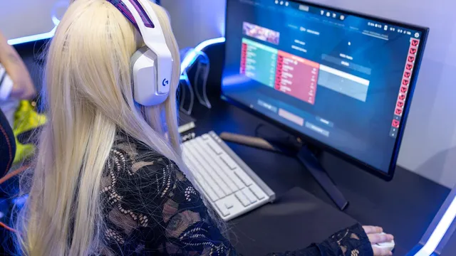 Jovem de cabelo louro comprido com auscultadores brancos a jogar num computador, com o ecrã e o teclado iluminados por luz azul