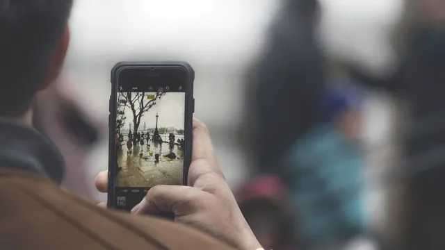 Pessoa de costas a fotografar com o telemóvel uma rua molhada à beira-rio, com a imagem visível no ecrã