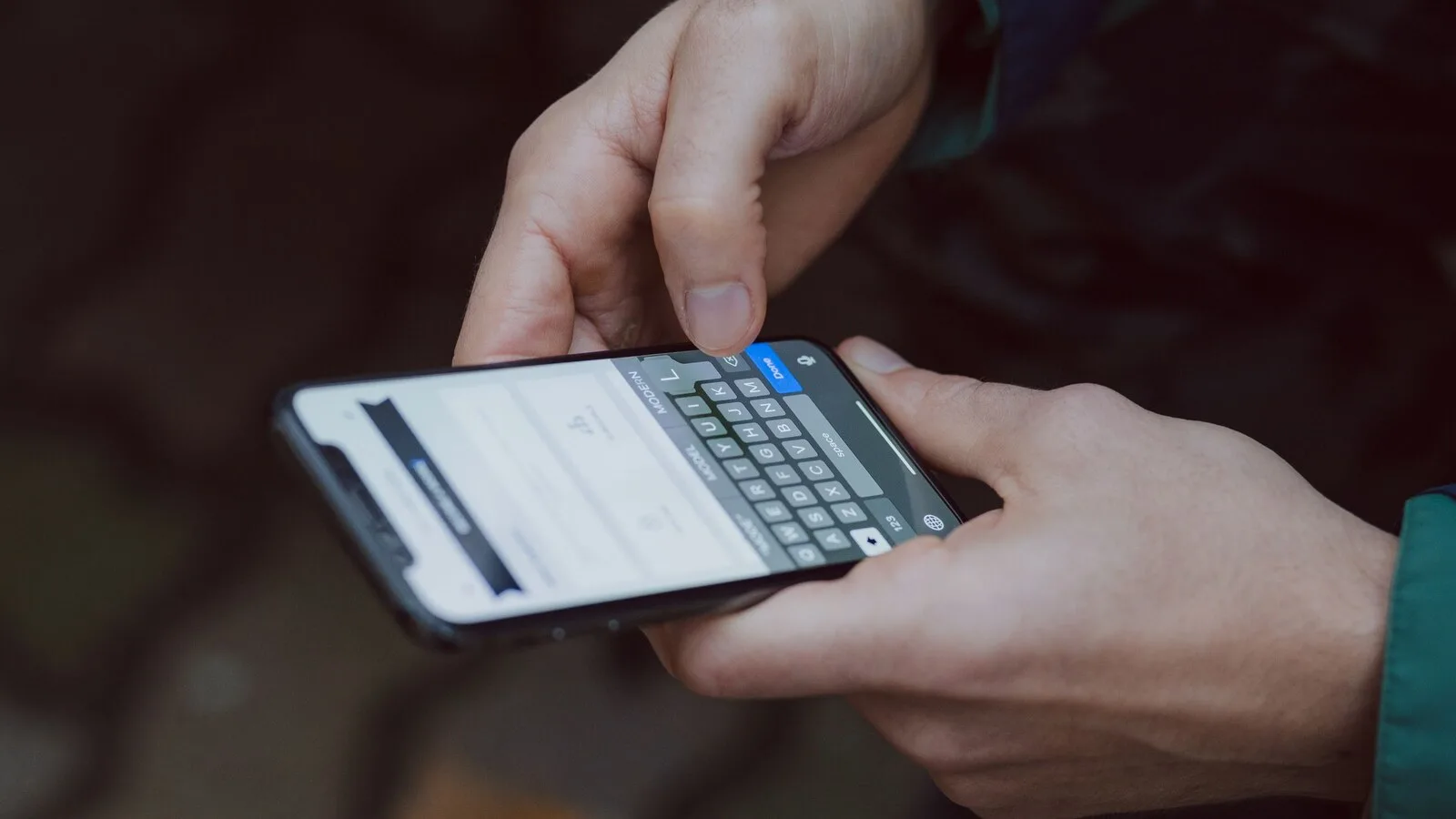 Mãos a escrever num telemóvel com o teclado aberto no ecrã, ao ar livre e com fundo desfocado