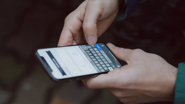 Mãos a escrever num telemóvel com o teclado aberto no ecrã, ao ar livre e com fundo desfocado