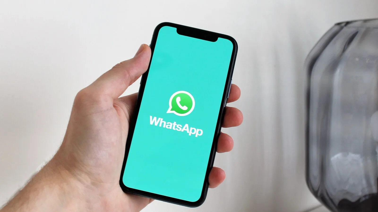 Mão a segurar um iPhone com o ecrã de arranque do WhatsApp, verde-água com o logótipo ao centro
