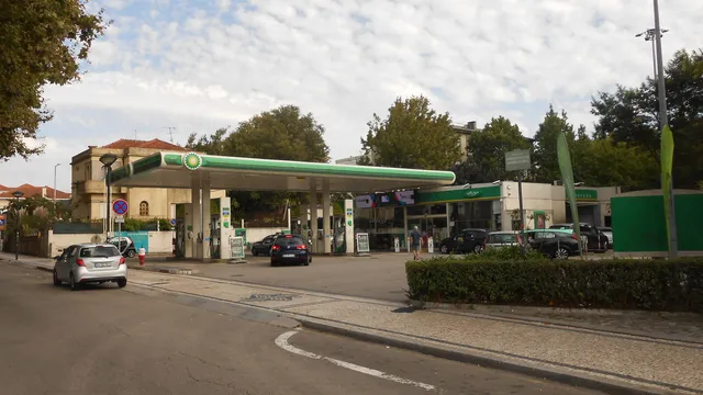 Posto de abastecimento com cobertura verde e branca numa rua arborizada, com carros a abastecer e um carro prateado a passar na estrada
