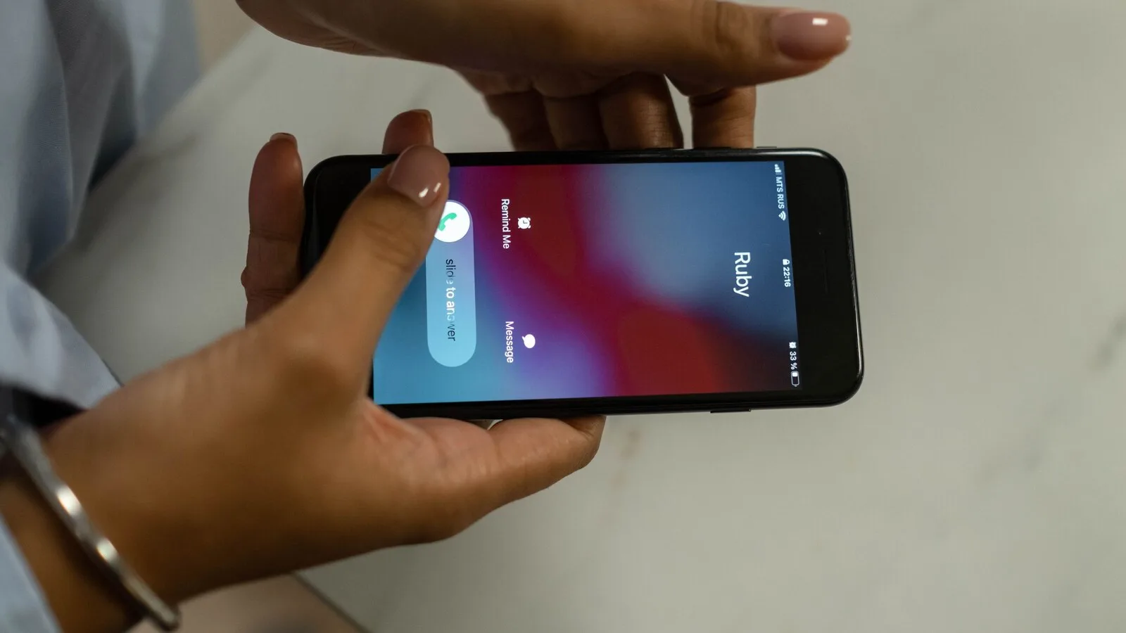 Duas mãos seguram um iPhone deitado sobre uma mesa de mármore, com o ecrã a mostrar uma chamada a chegar