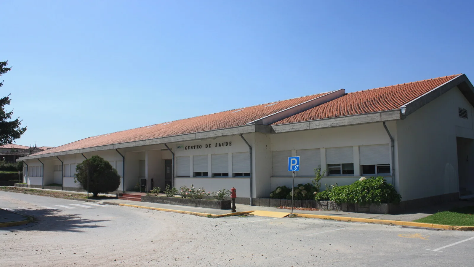 Edifício térreo de um centro de saúde com telhado de telha vermelha, letreiro Centro de Saúde na fachada e lugar de estacionamento para pessoas com mobilidade reduzida