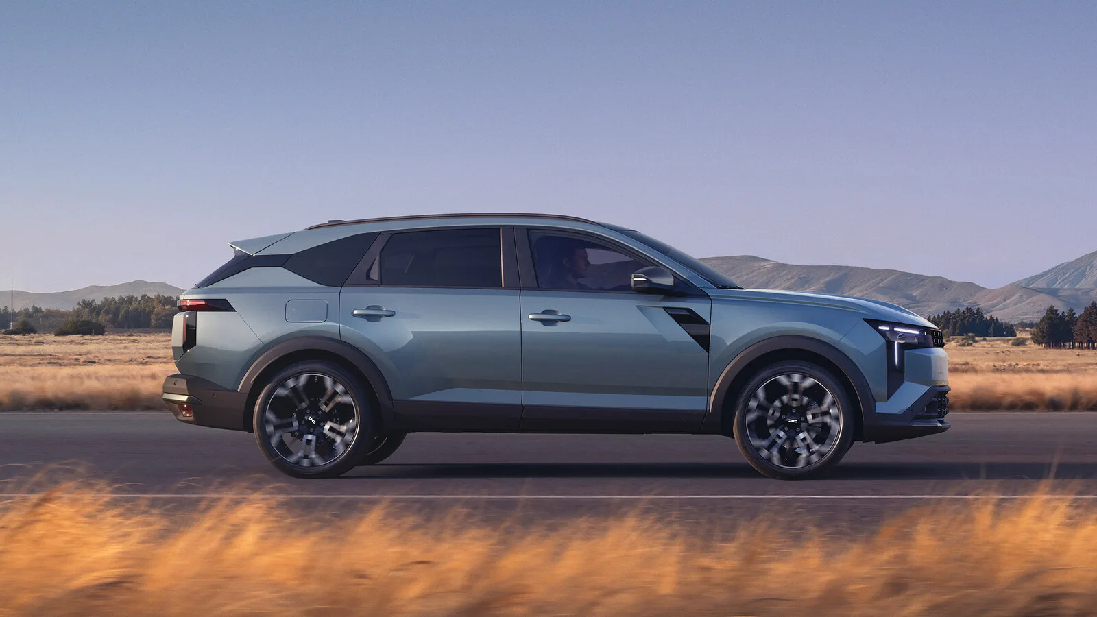 Dacia Striker em azul acinzentado visto de lado a circular numa estrada, com erva seca e montanhas ao fundo