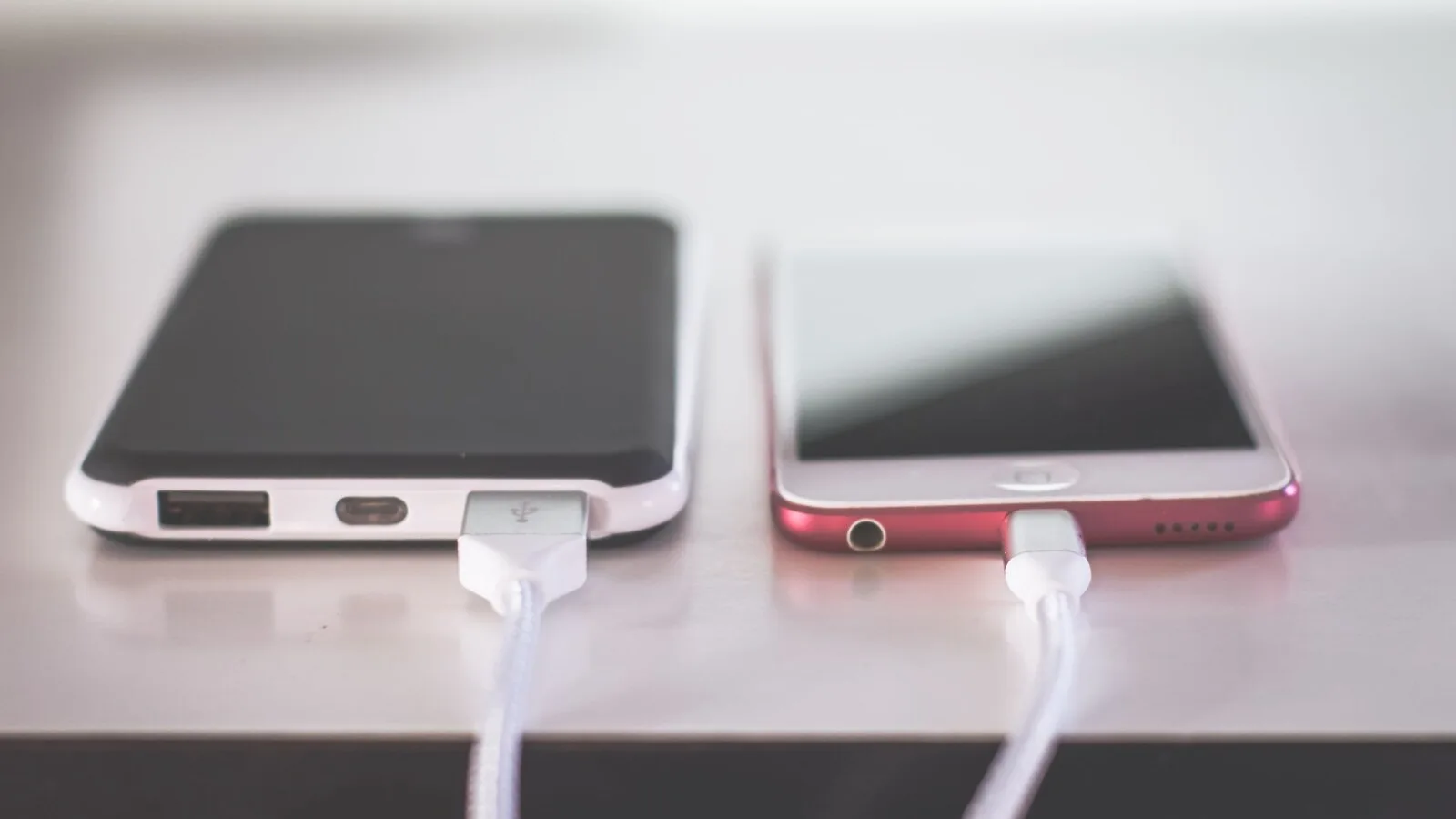Um telemóvel cor-de-rosa e uma bateria externa preta e branca, lado a lado sobre uma mesa, ligados a cabos de carregamento brancos